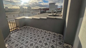 Een balkon met patroontegels, een metalen hekwerk en uitzicht op omliggende gebouwen onder een lichtbewolkte lucht.