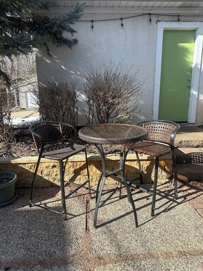 Une terrasse extérieure avec des chaises en métal et une table ronde, une porte verte et une guirlande lumineuse.
