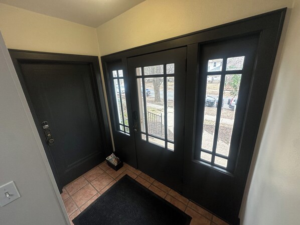 Un hall d’entrée aux couleurs sombres, doté d’un sol carrelé, d’un paillasson noir et d’une porte avec une fenêtre.