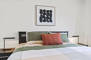 Une chambre à coucher bien rangée, avec un lit, des oreillers, une table de chevet et une œuvre d’art accrochée au mur.