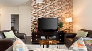 A living room with a brick wall, a flat-screen TV, leather sofas, and a coffee table. - Trailside Charm | TART Trail | Downtown | (Traverse City)