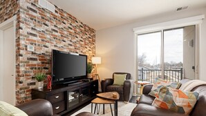 A living room with a brick wall, leather sofas, a TV, and a balcony with a view. - Trailside Charm | TART Trail | Downtown | (Traverse City)