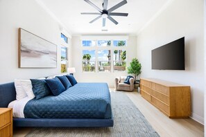 Une chambre à coucher avec un lit, une télévision, un ventilateur de plafond et une fenêtre donnant sur des palmiers.