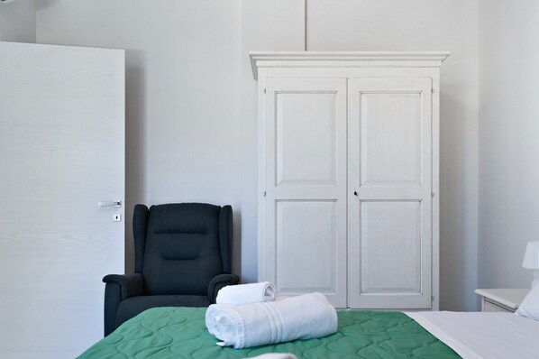 A hotel room with a green bed, a black chair, a white wardrobe, and a white bedside table with a lamp. - Renoir Apartment & Parking - Lecce Selection (Lecce)
