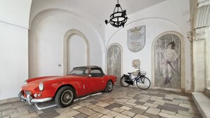 A red vintage car parked in a room with a checkered floor, a bicycle, and classical wall art.