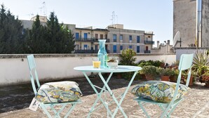 A rooftop terrace with two turquoise chairs, a small round table, and a vase.