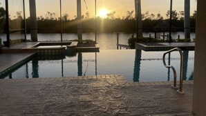 Una piscina infinita con vista al atardecer sobre una masa de agua, rodeada de palmeras y una estructura de vidrio.