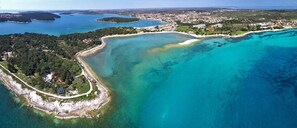 Vista aerea di una zona costiera con un'ampia baia, una piccola penisola con edifici e una fitta area boschiva.