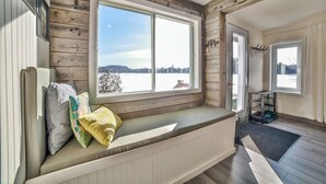 A room with a large window, wooden walls, a built-in bench with cushions, and a door leading to another room.