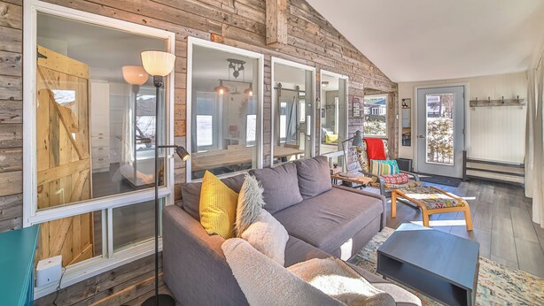 A cozy living room with a gray sofa, large windows, and wooden walls.