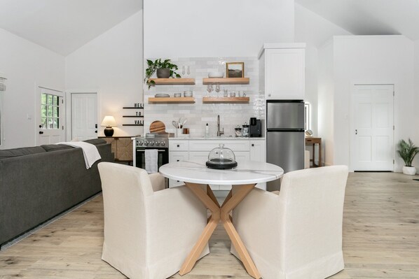 A modern kitchen with white cabinets, a built-in oven, and a stainless steel refrigerator. There is a dining area with a white table and two chairs.