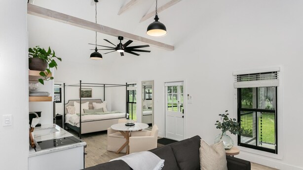 A modern living space with a central seating area, a dining table, and a kitchen area. There is a ceiling fan, a window with blinds, and a view of the outdoors. - Oak Valley | King Bed, Sun Deck & Outdoor Shower, Full Kitchen, Fire-pit (Fredericksburg)
