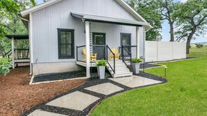 A single-story house with a covered porch, a small front yard with stepping stones, and a fenced yard. - Oak Valley | King Bed, Sun Deck & Outdoor Shower, Full Kitchen, Fire-pit (Fredericksburg)