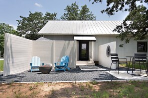 A small building with a metal roof, a door, and a window. There are two blue Adirondack chairs, a black metal table, and a large planter with rocks.