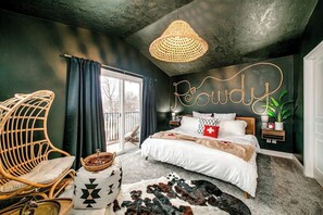 Ein Schlafzimmer mit einem Bett, einem Korbstuhl, einem Nachttisch, einer Wandlampe und einem großen Fenster.