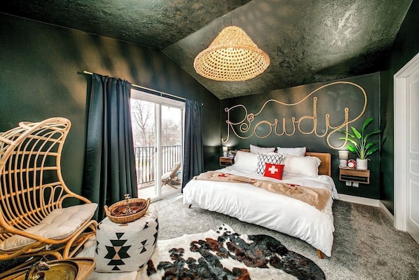 A bedroom with a bed, a wicker chair, a bedside table, a wall-mounted lamp, and a large window.