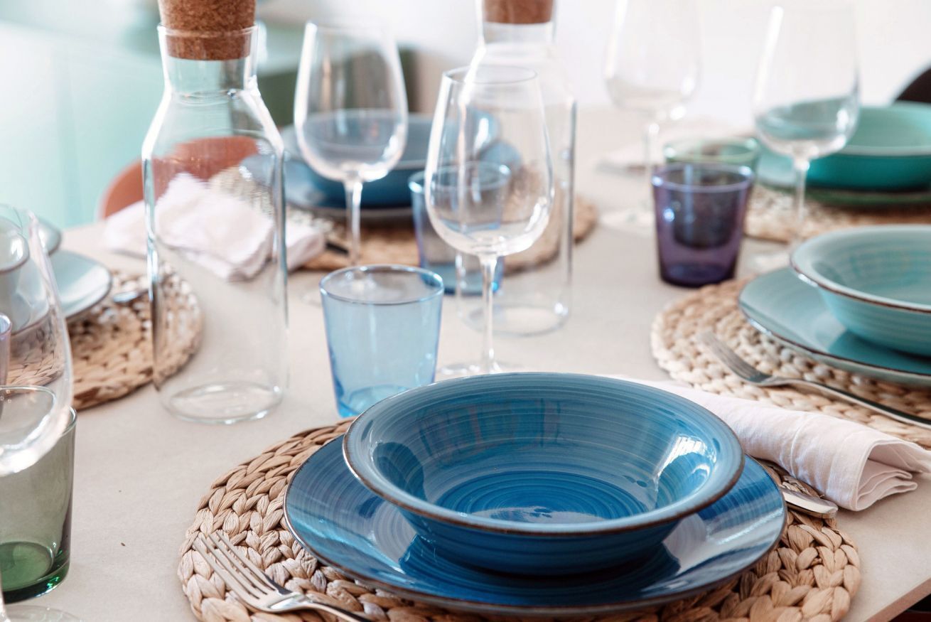 A table set with blue plates, clear glasses, and a corked bottle.
