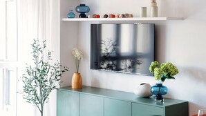 A modern living room with a flat-screen TV mounted on the wall, a teal cabinet below, and decorative items on a shelf above. - Como Hills - MyHomeinComo (Como)