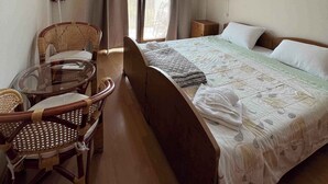 Une chambre d’hôtel comprenant un lit, une table en verre avec des chaises, une fenêtre avec des rideaux, un climatiseur fixé au mur et une petite table de chevet.