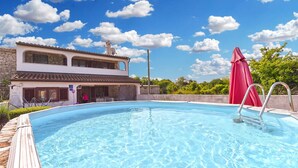 A swimming pool with a two-story building in the background, a red umbrella, and clear blue skies.