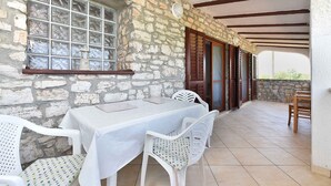 A stone-walled outdoor patio with a table and chairs, a window with a metal grate, and wooden doors.