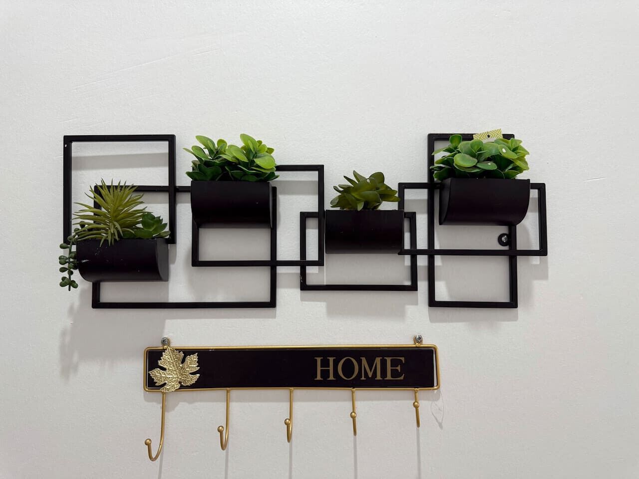 Un élément de décoration murale composé de quatre cadres carrés noirs, chacun contenant une plante. En dessous, une plaque noire portant l’inscription « HOME » en lettres dorées est suspendue à une tige métallique de couleur or.