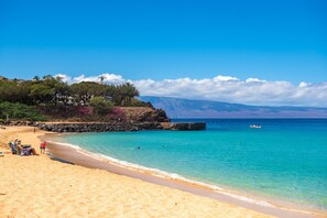 Une plage bordée d’une eau turquoise et limpide, un brise-lames rocheux et, au loin, une chaîne de montagnes.