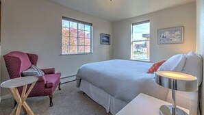 Een slaapkamer met een groot bed, twee ramen, een stoel, een klein tafeltje en een lamp.