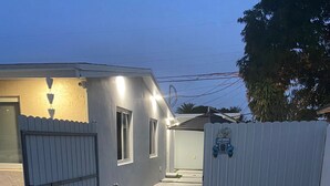 Une allée fermée par un portail mène à une maison entourée d’une clôture en bois peinte en blanc, et un panneau indique « Bienvenue ».