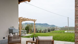 Une terrasse extérieure avec deux fauteuils en osier, une table en bois et une bouteille de vin. Une pergola offre de l’ombre sur l’espace détente.