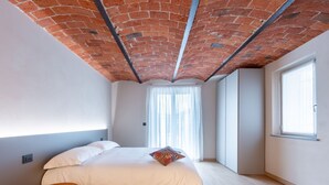 Une chambre moderne avec un plafond en briques, un grand lit avec une literie blanche, une petite table de chevet en bois et une fenêtre ornée de rideaux légers.