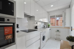 A modern kitchen with a built-in oven, microwave, and washing machine, a window with a view of buildings, and a potted plant.
