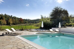 Une piscine entourée de chaises longues et de parasols, bordée d’une terrasse en pierre et offrant une vue sur une colline boisée.