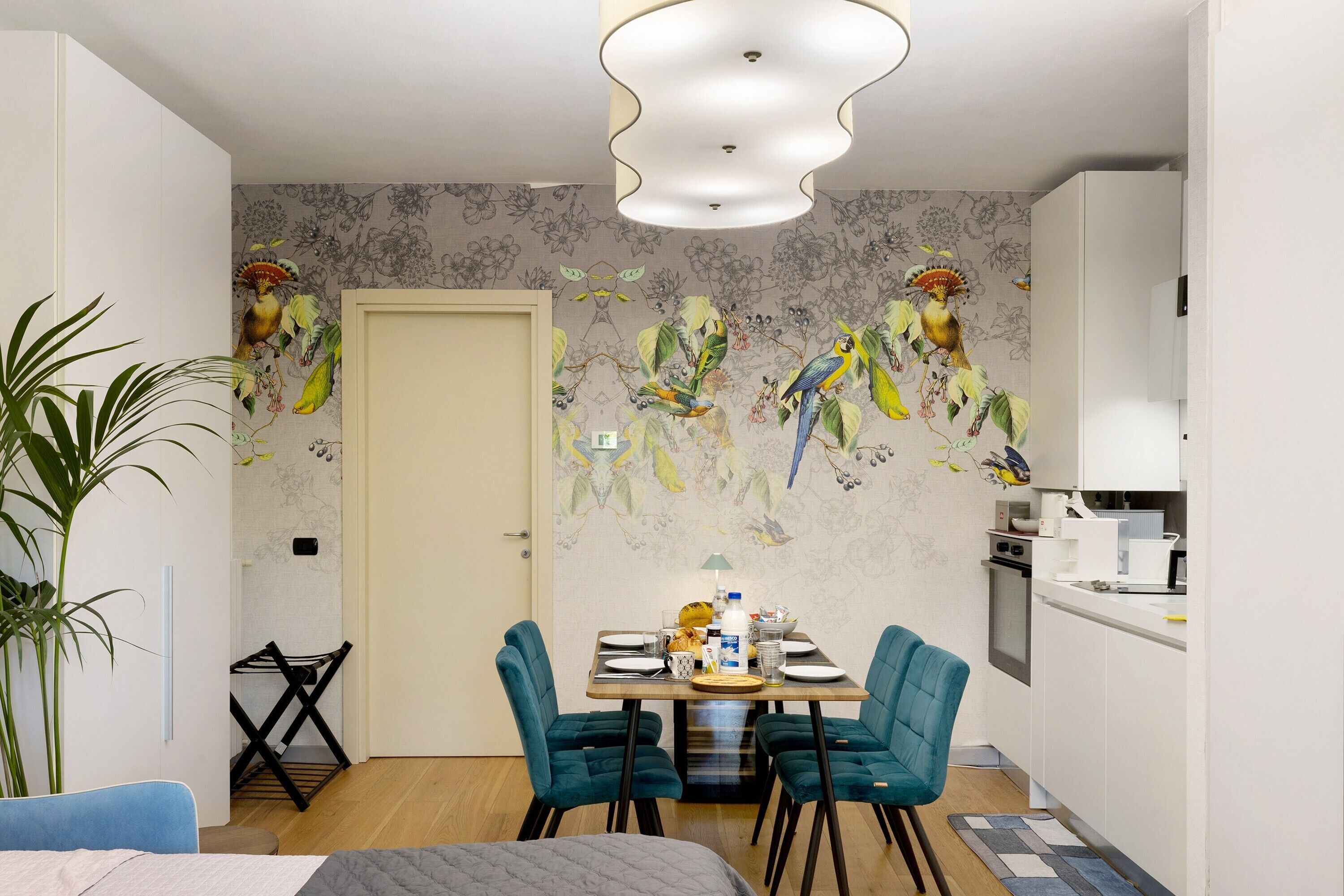 A dining area with a table set for four, two teal chairs, a black chair, a white door, and a kitchen area with white cabinets.