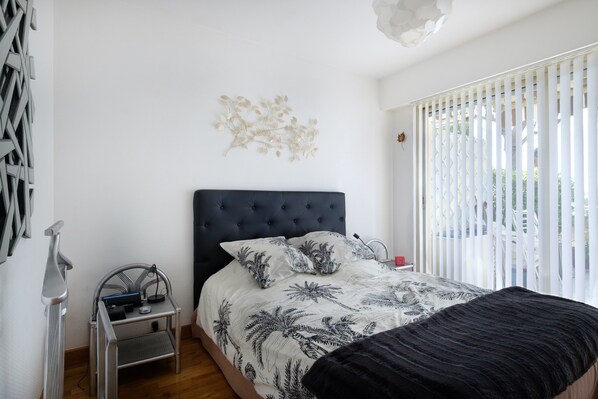 A bedroom with a bed, a small table, a wall-mounted light fixture, and a window with blinds.