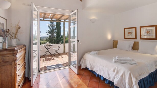 Un dormitorio con cama, cómoda, balcón con mesa y sillas, y vista a árboles.