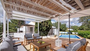 Un patio exterior con mobiliario de madera, una piscina y vistas del paisaje circundante.