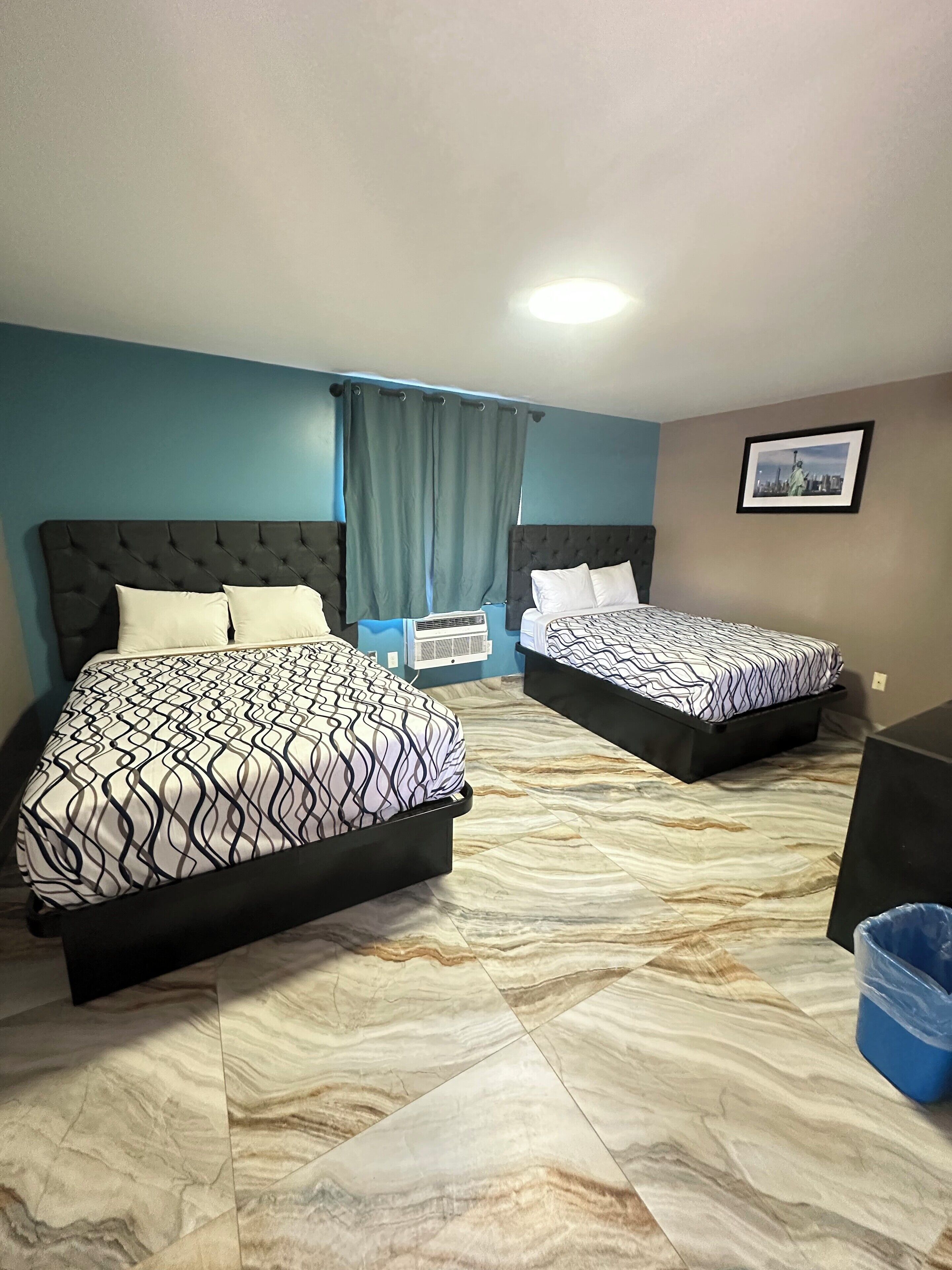 A hotel room with two beds, a blue wall, a framed picture, and a blue trash bin.