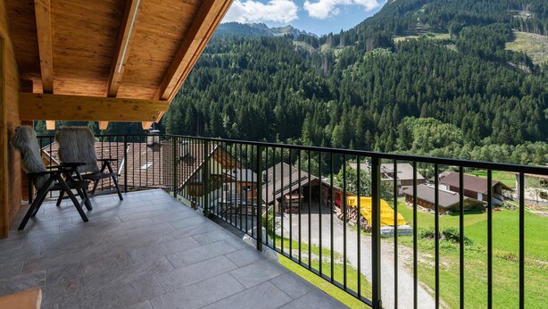 A wooden balcony with chairs, a view of a mountainous landscape, and a building with a yellow tent in the background. - Apartment 'Lodge 3' with Mountain View, Shared Terrace and Wi-Fi (Colle Isarco)