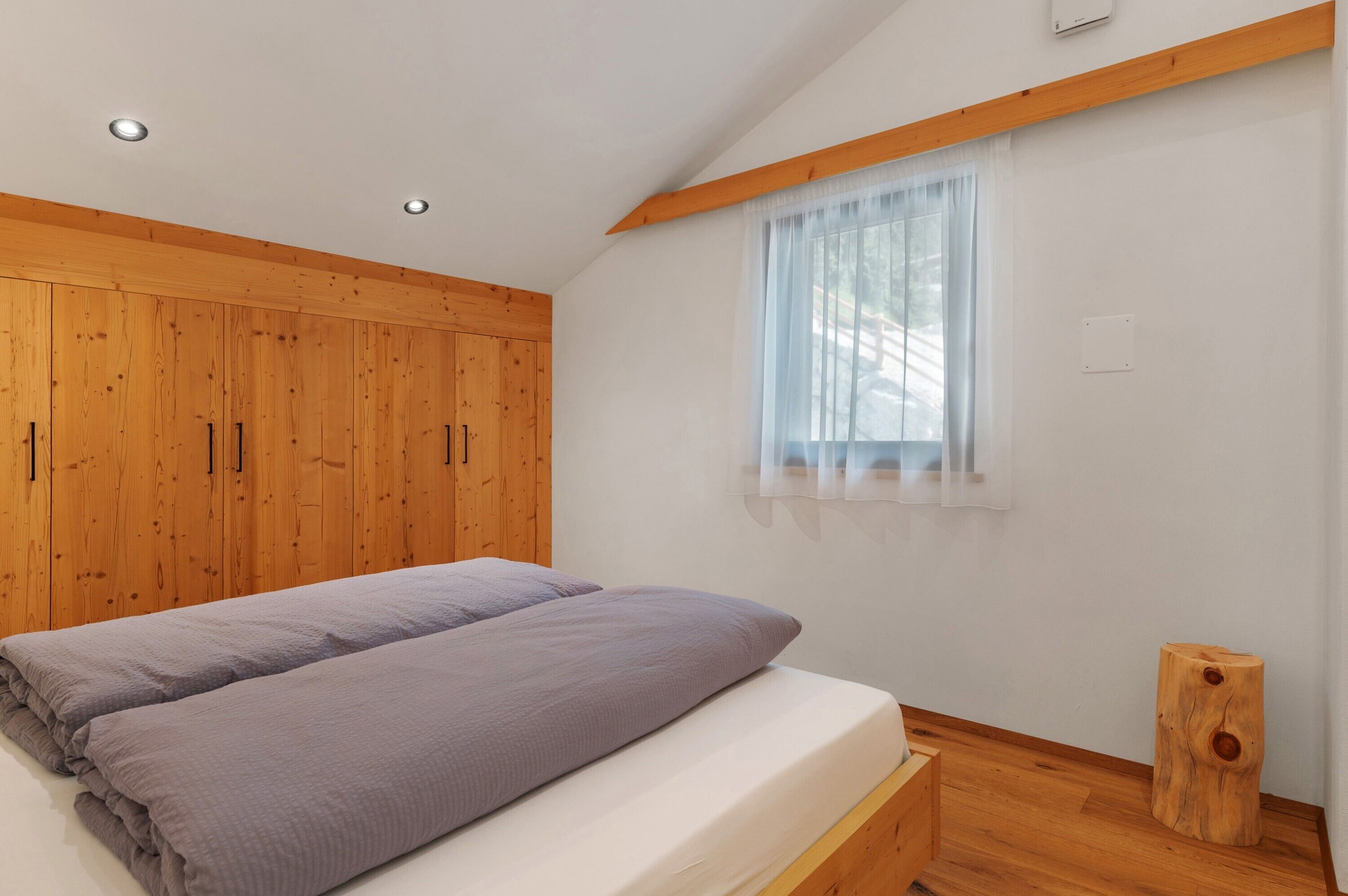 Une chambre à coucher avec un lit, une armoire en bois, une fenêtre avec des rideaux légers et un tabouret en bois.