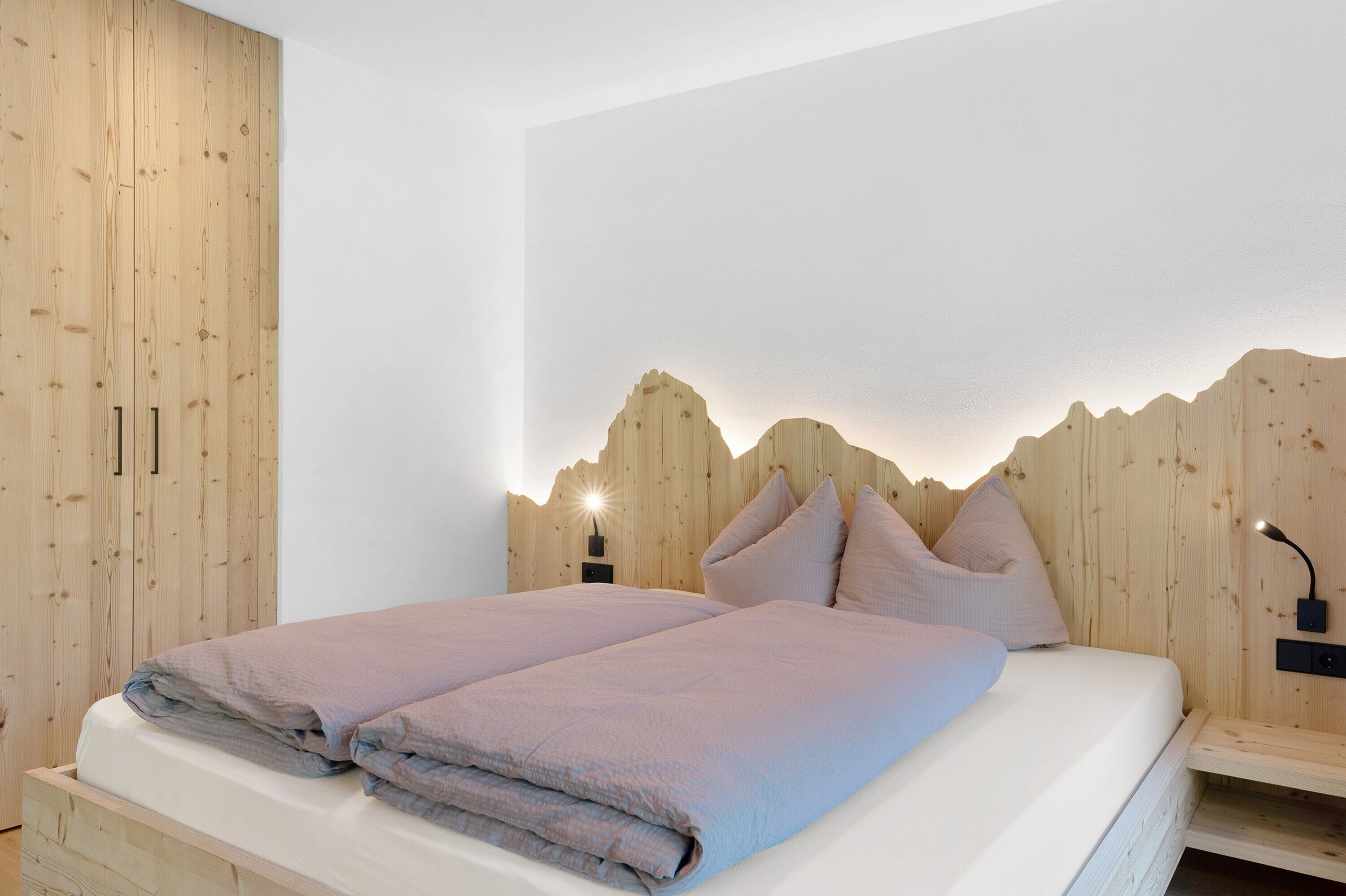A bedroom with a wooden headboard, two beds, bedside tables, and a wall-mounted lamp.
