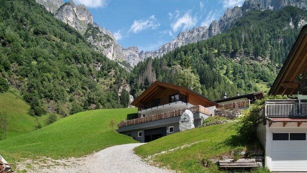 Un chalet con un balcone, un muro in pietra e uno sfondo montuoso.