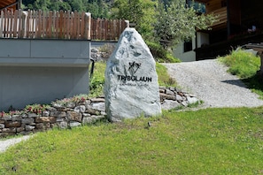 A stone sign with 'TRIBULAUN' and a mountain symbol, set against a backdrop of a wooden fence, stone wall, and a building with a sloped roof.
