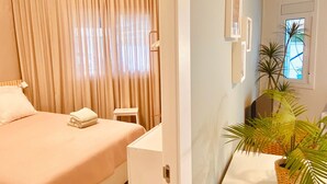 A hotel room with a bed, a desk, a chair, a small table, a potted plant, and a window.