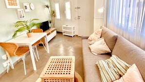 A living room with a sofa, a TV, a dining table, chairs, a small wooden stool, and a ceiling fan.