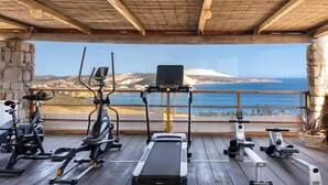 Een overdekte buitenfitnessruimte met trainingsapparatuur, met uitzicht op een prachtig zee- en heuvellandschap.