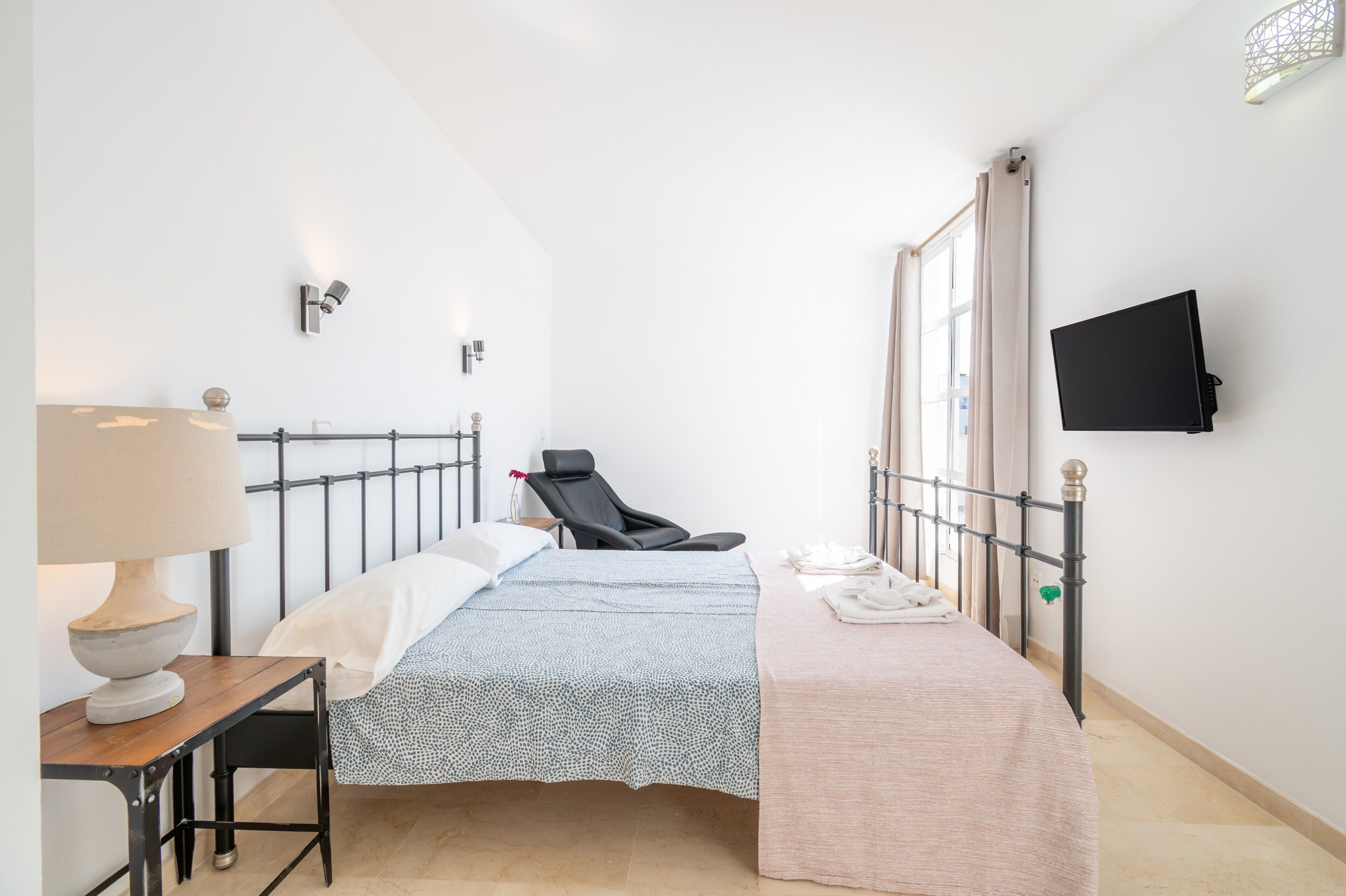 Une chambre à coucher comprenant un lit, une chaise, une table avec une lampe, un téléviseur à écran plat et une fenêtre avec des rideaux.