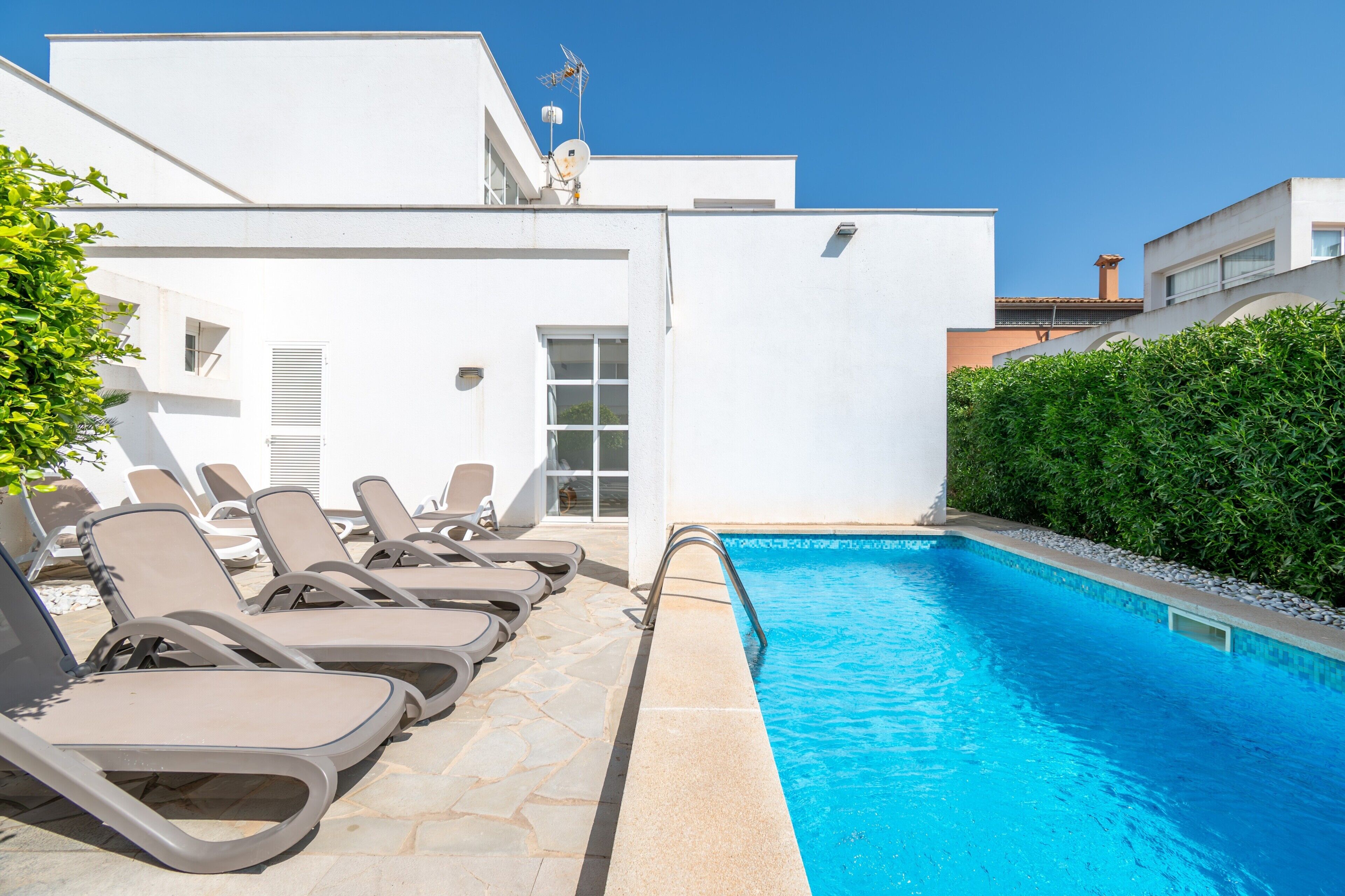 A swimming pool with lounge chairs, a white building, and a hedge.