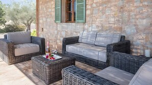 An outdoor seating area with wicker furniture, a table with a bottle and glasses, and a stone wall with green shutters. - Country House 'Calma' with Mountain View, Private Pool and Wi-Fi (Illes Balears)