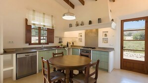 A kitchen with a dining table, chairs, a dishwasher, a stove, and a window with blinds. - Country House 'Calma' with Mountain View, Private Pool and Wi-Fi (Illes Balears)